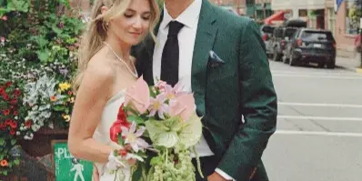 A bride and groom posing on a street in a mountain town with a bouquet and green suit.