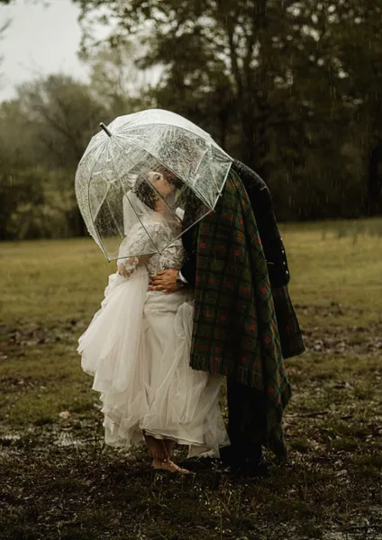 A bride and groom sharing a kiss under a clear umbrella in a misty, wooded field.