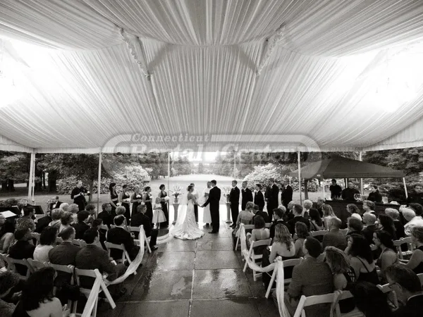 A black and white photograph of a wedding ceremony held under a large white tent with rows of chairs and guests.