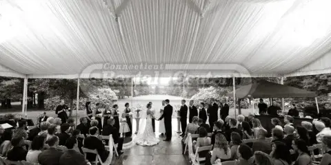 A black and white photograph of a wedding ceremony held under a large white tent with rows of chairs and guests.