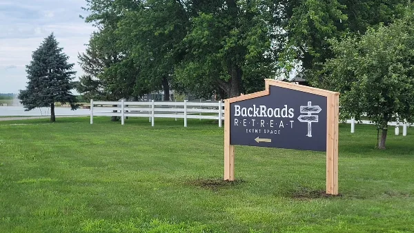 Exterior view of the BackRoads Retreat Event Space sign in a grassy area with a white fence and trees in the background.