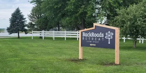 Exterior view of the BackRoads Retreat Event Space sign in a grassy area with a white fence and trees in the background.