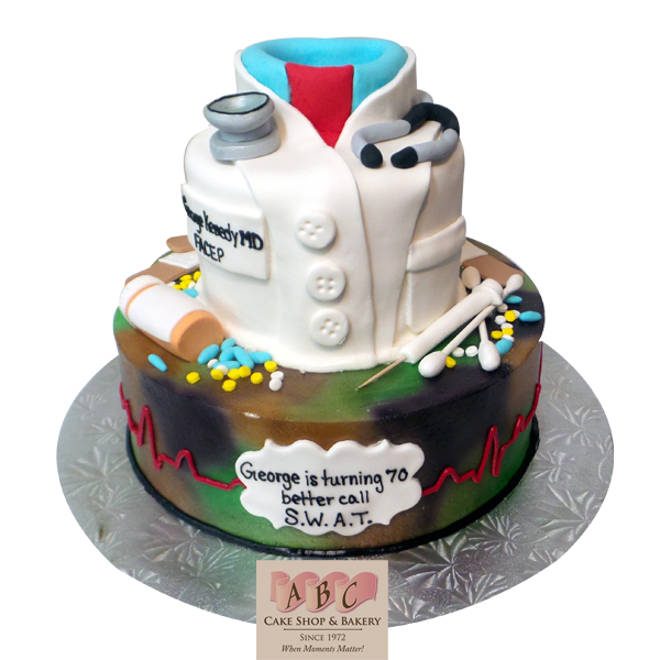 A two-tier custom cake shaped like a doctor's coat with a stethoscope and medical tools, sitting on a silver cake stand with a branded card.