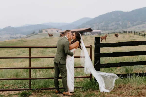 A bride and groom kissing in a grassy field with a wooden fence and mountains in the background.