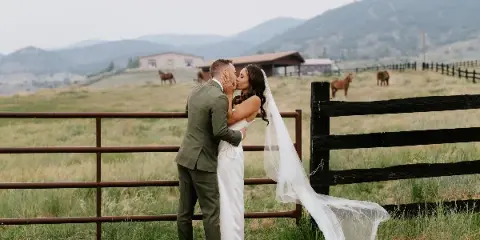 A bride and groom kissing in a grassy field with a wooden fence and mountains in the background.