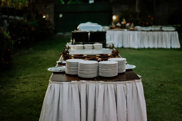 An outdoor catering setup featuring a table with stacks of white plates and buffet equipment.