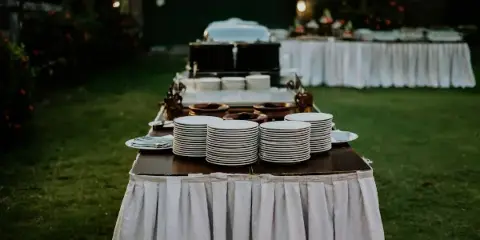 An outdoor catering setup featuring a table with stacks of white plates and buffet equipment.