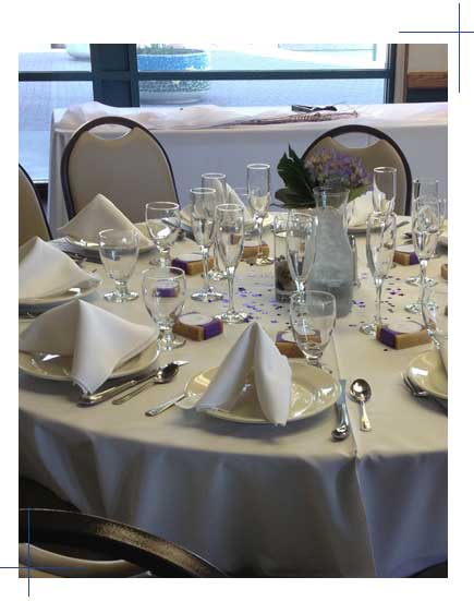 A formally set catering table with white linens, glassware, folded napkins, and a floral centerpiece.