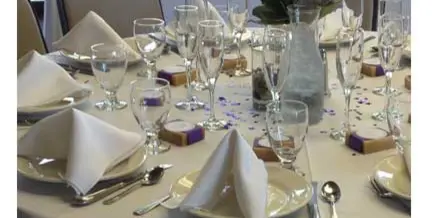 A formally set catering table with white linens, glassware, folded napkins, and a floral centerpiece.