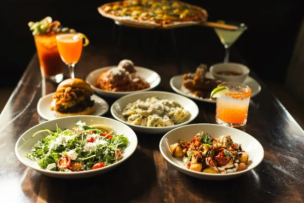 A table spread featuring multiple Italian dishes including a Caesar salad, pasta, a burger, and a pizza in the background.