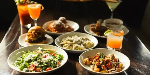 A table spread featuring multiple Italian dishes including a Caesar salad, pasta, a burger, and a pizza in the background.