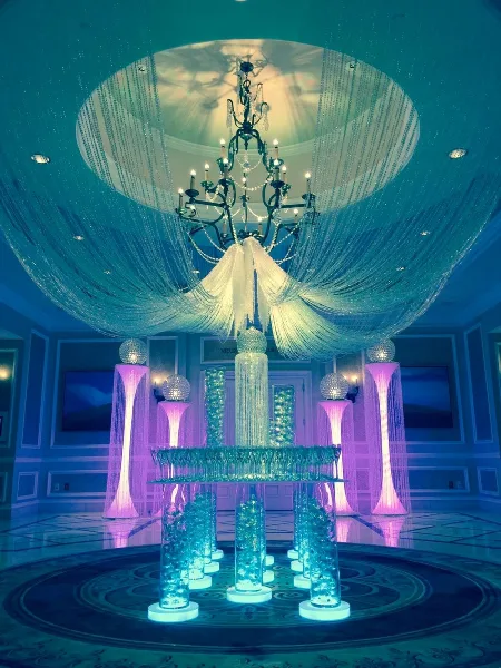 An elaborate event decor setup featuring a large crystal chandelier, a 360-degree drape canopy, and illuminated glass pedestals in a ballroom setting.