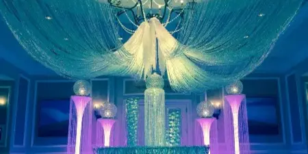 An elaborate event decor setup featuring a large crystal chandelier, a 360-degree drape canopy, and illuminated glass pedestals in a ballroom setting.