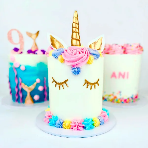 A white unicorn-themed cake with a gold horn, ears, and closed eyes, decorated with colorful frosting flowers.