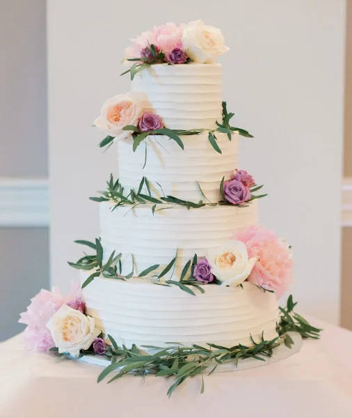 A four-tier white wedding cake decorated with pink and purple roses and greenery.