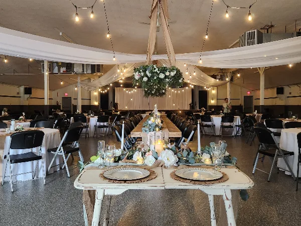 Styled wedding reception room with long tables, white linens, candles, floral centerpieces, hanging floral installation, and string lights.