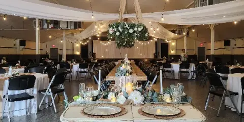 Styled wedding reception room with long tables, white linens, candles, floral centerpieces, hanging floral installation, and string lights.
