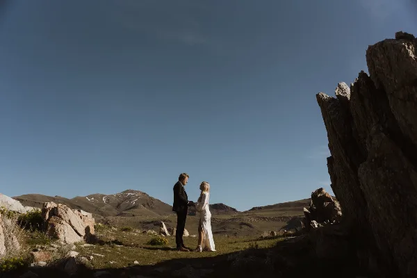 A couple in wedding attire standing together in a mountainous landscape with rocky outcrops and a clear blue sky.