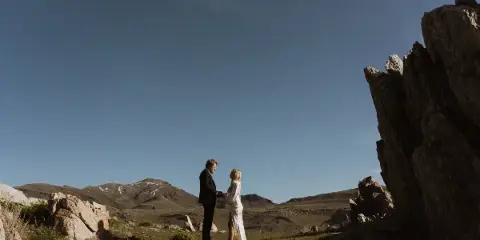 A couple in wedding attire standing together in a mountainous landscape with rocky outcrops and a clear blue sky.