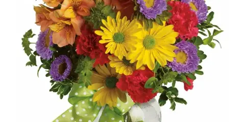 A colorful mixed bouquet of flowers including yellow daisies, red carnations, and purple chrysanthemums in a clear glass vase with a green polka-dot ribbon.
