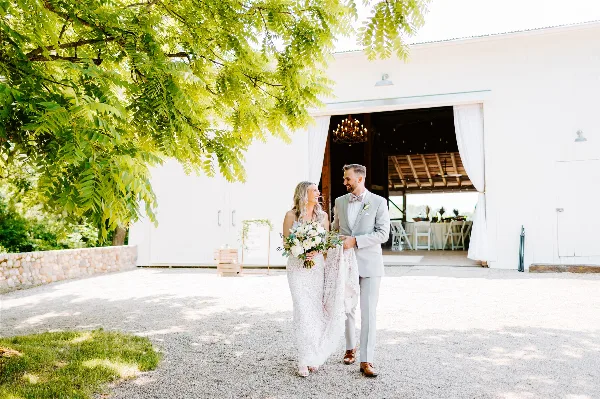 A bride and groom standing outside a white wedding barn venue with a chandelier visible inside.