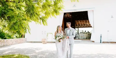 A bride and groom standing outside a white wedding barn venue with a chandelier visible inside.
