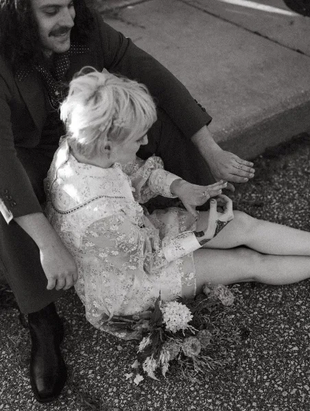 A black and white documentary-style photograph of a bride and groom sitting on the ground, sharing an intimate moment with the bride's bouquet nearby.
