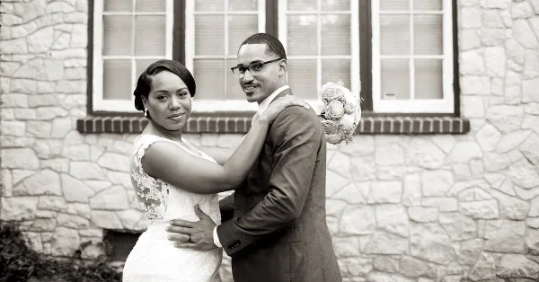 A black and white photograph of a bride and groom embracing in front of a stone building.