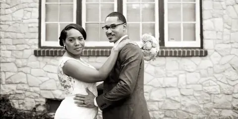 A black and white photograph of a bride and groom embracing in front of a stone building.