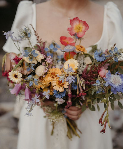 A person in a white dress holding a vibrant, mixed bouquet of seasonal flowers including roses, cosmos, and blue cornflowers.
