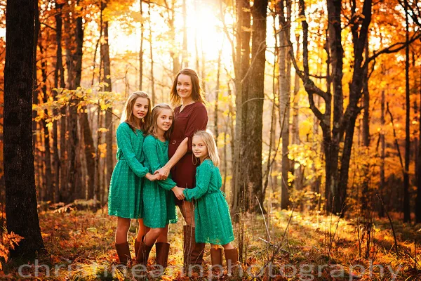 A mother and four young girls in green dresses posing together in a sunlit autumn forest.