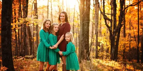 A mother and four young girls in green dresses posing together in a sunlit autumn forest.