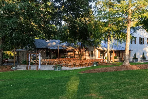 Outdoor wedding ceremony setup with wooden arch and rows of chairs in front of a rustic barn-style venue building surrounded by trees and lawn.