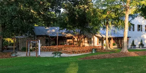 Outdoor wedding ceremony setup with wooden arch and rows of chairs in front of a rustic barn-style venue building surrounded by trees and lawn.