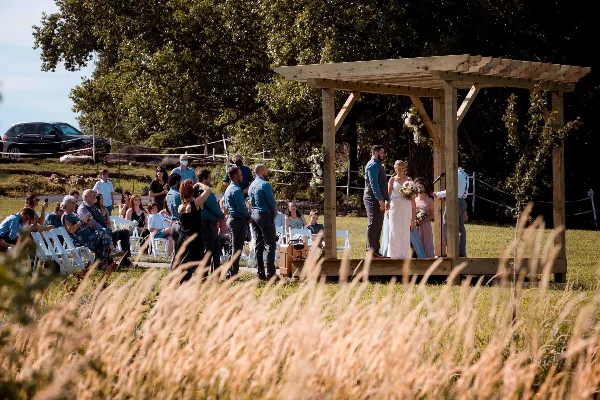 A bride and groom standing under a wooden pergola during an outdoor wedding ceremony with seated guests.