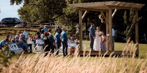 A bride and groom standing under a wooden pergola during an outdoor wedding ceremony with seated guests.