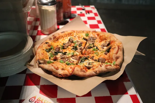 A wood-fired pizza with mushrooms and greens served on parchment paper on a red-and-white checkered catering table.