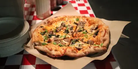 A wood-fired pizza with mushrooms and greens served on parchment paper on a red-and-white checkered catering table.