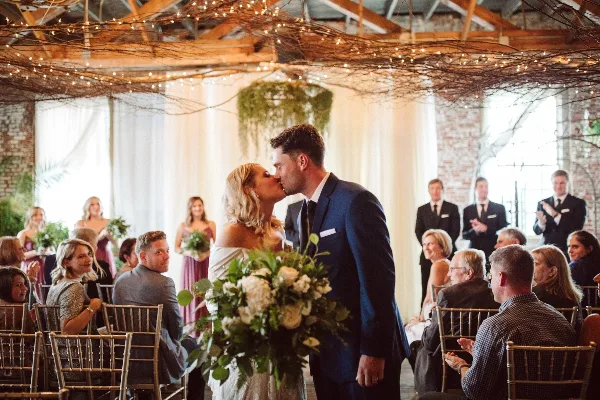 A bride and groom share a kiss during a wedding reception in a venue with exposed brick walls and string lights.