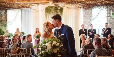 A bride and groom share a kiss during a wedding reception in a venue with exposed brick walls and string lights.