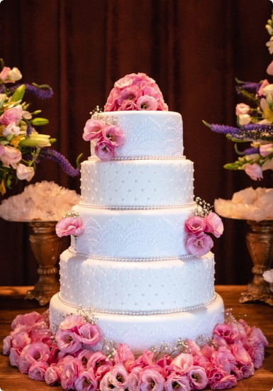 A four-tier white wedding cake decorated with pink roses and pearl trim, displayed on a table with floral arrangements.
