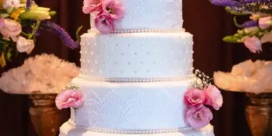 A four-tier white wedding cake decorated with pink roses and pearl trim, displayed on a table with floral arrangements.
