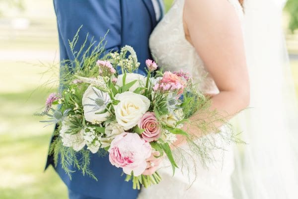 A close-up of a bride and groom holding a lush, garden-style wedding bouquet featuring pink and white roses, thistles, and greenery.