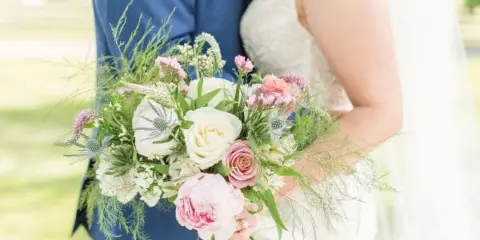 A close-up of a bride and groom holding a lush, garden-style wedding bouquet featuring pink and white roses, thistles, and greenery.