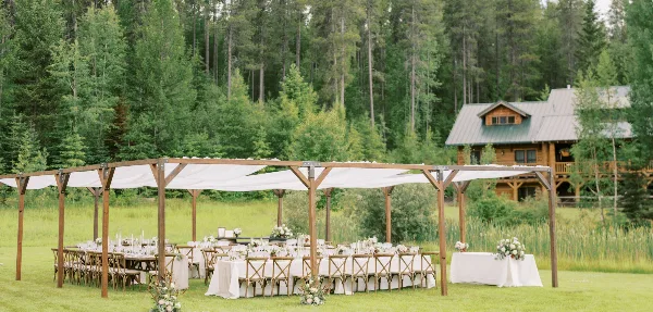 Outdoor wedding reception setup with a wooden pergola, white draping, set tables, and floral centerpieces in a grassy field with a forest and cabin in the background.