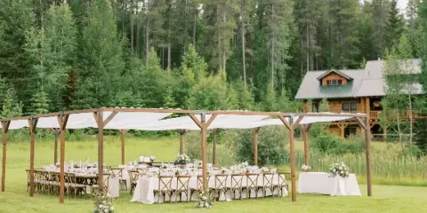 Outdoor wedding reception setup with a wooden pergola, white draping, set tables, and floral centerpieces in a grassy field with a forest and cabin in the background.