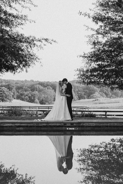 Black and white photo of a bride and groom embracing on a stone ledge by a pond, with trees and a field in the background.