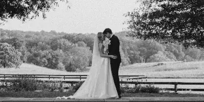 Black and white photo of a bride and groom embracing on a stone ledge by a pond, with trees and a field in the background.