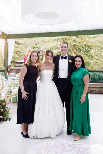 A bride and groom posing with two women in formal attire at a decorated wedding venue with floral arrangements and white drapery.