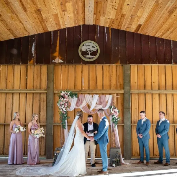 A bride and groom exchanging vows at a floral arch inside a wooden barn venue, flanked by bridesmaids and groomsmen.
